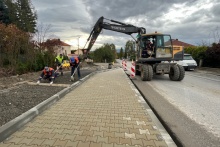 na drodze stoi koparka wykonująca wykopy wzdłuż chodnika, obok pracownicy budowlani w trakcie pracy 