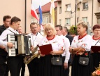 grupa ludzi śpiewająca przy kościele, tuż za nimi widnieje flaga polski. Jeden meżczyzna trzyma w rękachj akordeon 