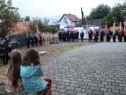 plac na którym odbywją się uroczystości jubileuszu, dwie dziwczynki zwrócone plecami patrzą na uroczystość, widać orkiestrę, MDP oraz strażaków i poczty sztandarowe