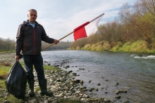 fot. nadesłane przez Stowarzyszenie  Przyjaciele Raby, na zdjęciu mężczyzna na brzegu rzeki, trzymający w jednej ręce flagę Polski, w drugiej worek ze śmieciami