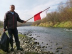 fot. nadesłane przez Stowarzyszenie  Przyjaciele Raby, na zdjęciu mężczyzna na brzegu rzeki, trzymający w jednej ręce flagę Polski, w drugiej worek ze śmieciami